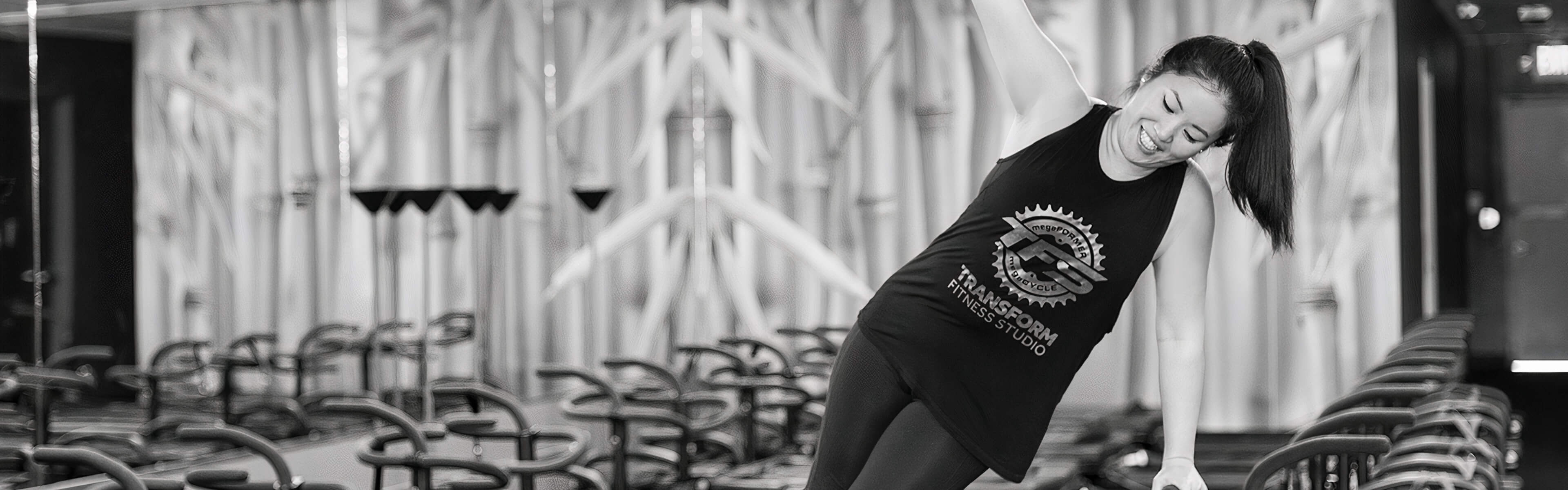 Andrea Ruelas smiling in a side plank in a Transform Fitness tank, Megaformers lined up behind her